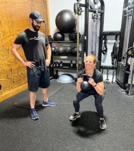 A person performing a kettlebell squat while a trainer observes in a gym setting
